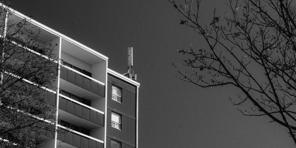 Modernes Apartmentgebäude mit mehreren Balkonen und kahlen Bäumen im Vordergrund, fotografiert bei Tageslicht.