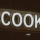 Nahaufnahme eines weißen Schilds mit dem Schriftzug „Cook“, von Jon Tyson fotografiert, geeignet für Gastronomie-Themen.