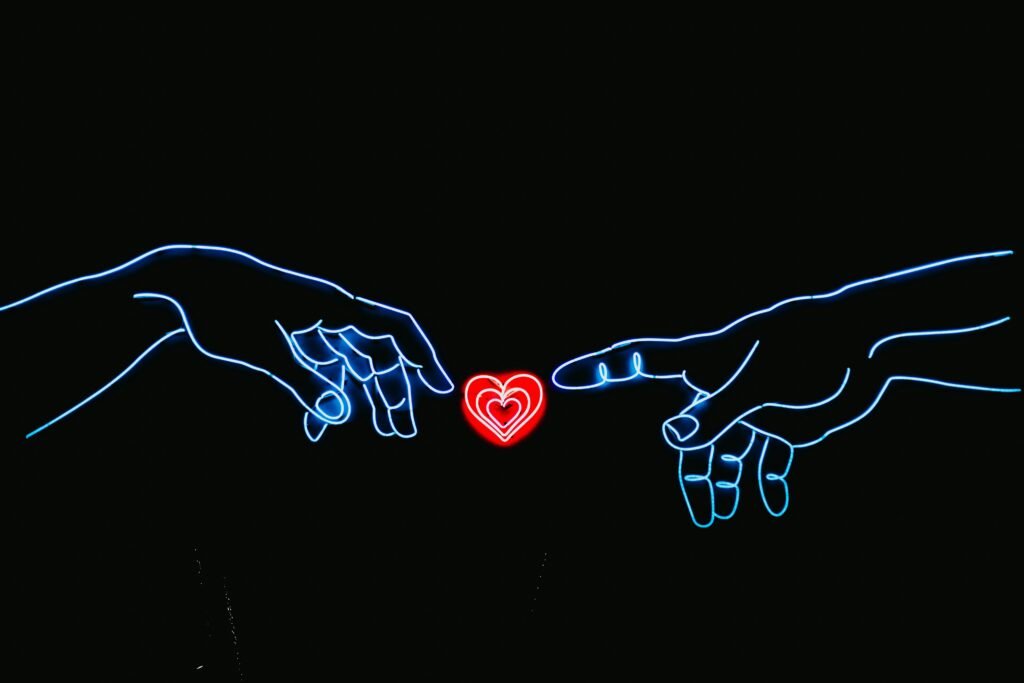 Zwei Hände nähern sich einem herzförmigen Neonlicht auf dunklem Hintergrund, rote und blaue Farbakzente erleuchten das Bild.
