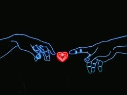 Zwei Hände nähern sich einem herzförmigen Neonlicht auf dunklem Hintergrund, rote und blaue Farbakzente erleuchten das Bild.