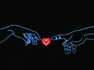Zwei Hände nähern sich einem herzförmigen Neonlicht auf dunklem Hintergrund, rote und blaue Farbakzente erleuchten das Bild.
