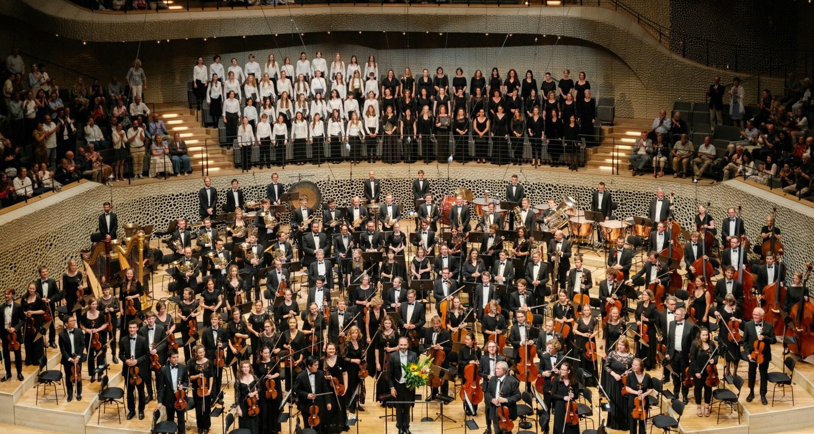 Ein Orchester und ein Chor führen zusammen ein Musikstück auf einer Konzertbühne vor, mit Sängern und Instrumenten.