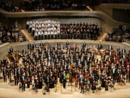 Ein Orchester und ein Chor führen zusammen ein Musikstück auf einer Konzertbühne vor, mit Sängern und Instrumenten.