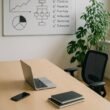 Modernes minimalistisches Büro mit großem Tisch, Laptop, Smartphone, Whiteboard mit Diagrammen und Pflanzen, helles natürliches Licht, ergonomischer Stuhl.