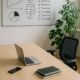 Modernes minimalistisches Büro mit großem Tisch, Laptop, Smartphone, Whiteboard mit Diagrammen und Pflanzen, helles natürliches Licht, ergonomischer Stuhl.