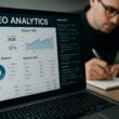 Nahaufnahme eines Laptops mit SEO-Dashboards, Charts, Grafiken und Code, im Hintergrund eine Person mit Brille beim Notieren, auf einem modernen Schreibtisch mit Smartphone und externen Festplatten.
