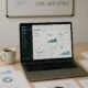 Moderner Arbeitsplatz mit Laptop, gedruckten Berichten, Lupe, Whiteboard mit SEO-Schlagwörtern und Kaffee, betont technische Optimierung und Analytics.