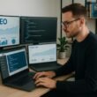 Professioneller männlicher Bloggger sitzend an modernem Schreibtisch mit mehreren Monitoren, die SEO-Daten, Code und Website-Performance anzeigen, in minimalistischer Büroumgebung