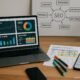 Moderner Arbeitsplatz mit Laptop, gedruckten Keyword-Daten, Whiteboard mit SEO-Diagrammen und grüner Pflanze