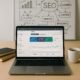 Arbeitsplatz mit Laptop, Notizbüchern und Whiteboard mit SEO-Skizzen, dargestellt auf einer modernen Arbeitsfläche