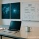 Moderner Arbeitsplatz in hellem Büro mit Laptop, Notizblock, Whiteboards, Digitalmaps und Infografiken, die Vernetzung und Technik thematisieren.