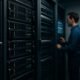 Nahaufnahme eines modernen Serverraums mit dunklen Serverracks, blinkenden LED-Leuchten und einem Techniker, der die Systeme auf einem digitalem Kontrollpanel überwacht.