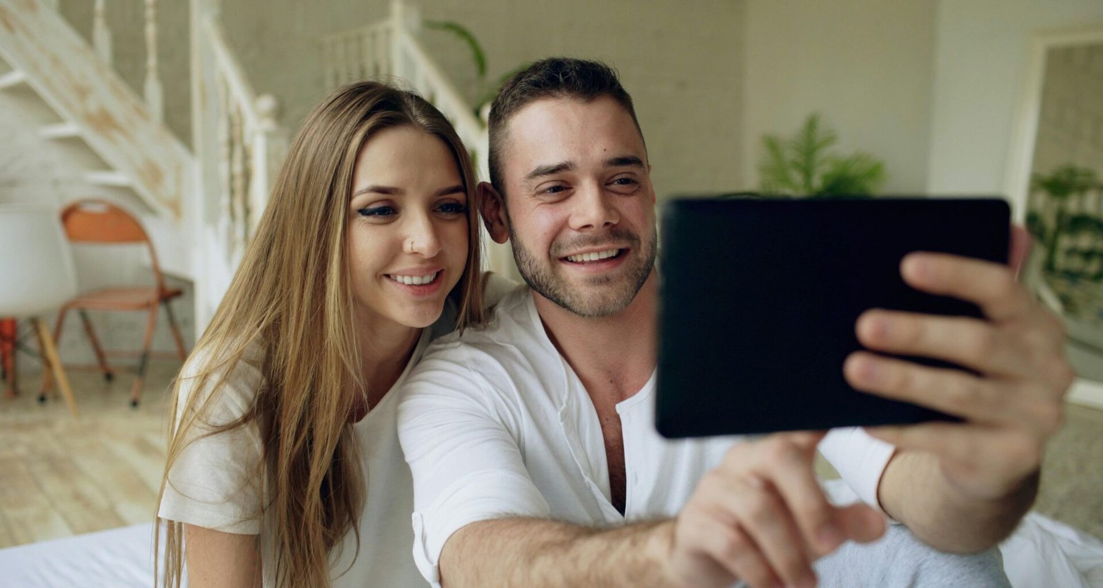 Ein Paar sitzt zusammen und nimmt ein Selfie mit einem Tablet auf, beide blicken in die Kamera des Geräts