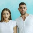 Ein Paar mit deutlich schockierten Gesichtern steht gemeinsam vor einem hellblauen Hintergrund im Studio.