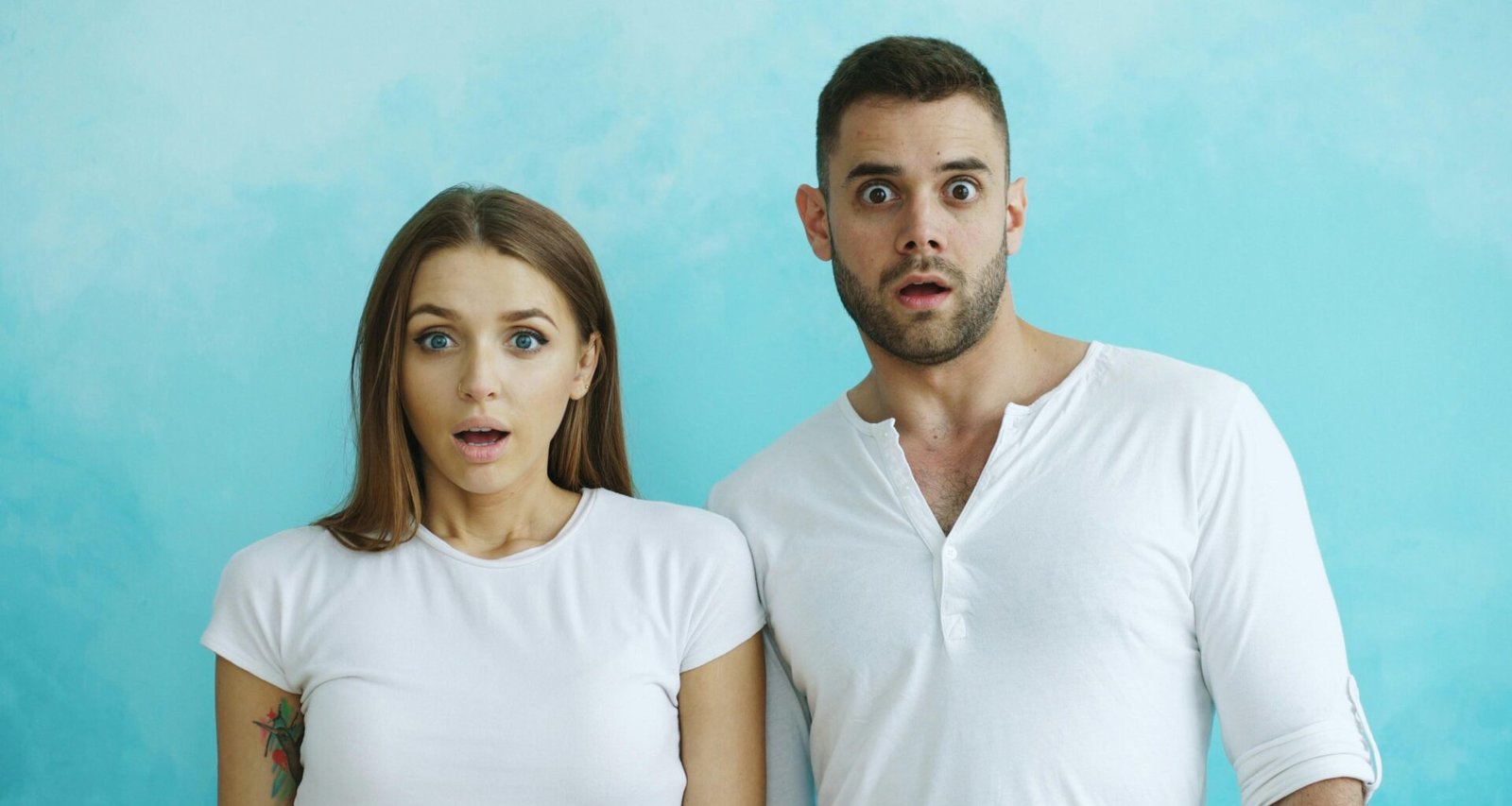 Ein Paar mit deutlich schockierten Gesichtern steht gemeinsam vor einem hellblauen Hintergrund im Studio.