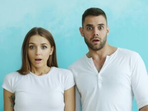 Ein Paar mit deutlich schockierten Gesichtern steht gemeinsam vor einem hellblauen Hintergrund im Studio.