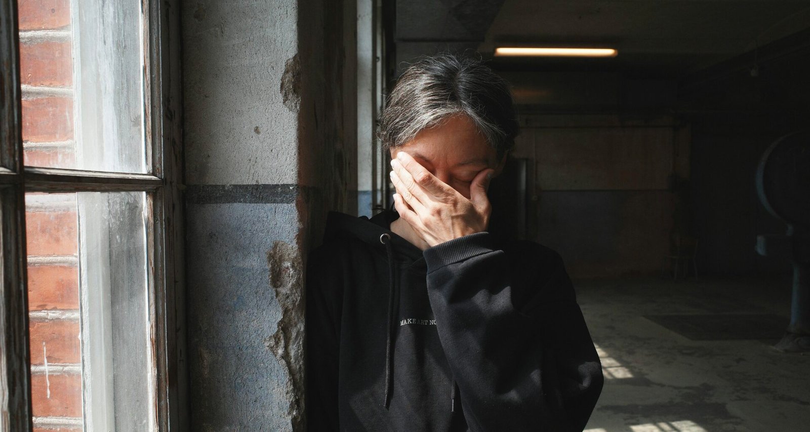 Person steht nahe einem Fenster und bedeckt das Gesicht mit der Hand, Tageslicht fällt herein, Innenraum, neutraler Hintergrund