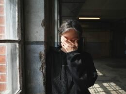 Person steht nahe einem Fenster und bedeckt das Gesicht mit der Hand, Tageslicht fällt herein, Innenraum, neutraler Hintergrund