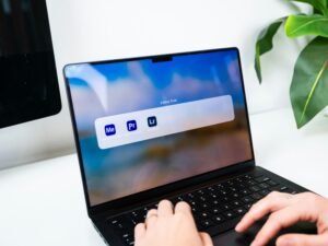 Person sitzt an einem Schreibtisch und tippt auf einem Laptop, auf dessen Bildschirm verschiedene Softwaresymbole zu sehen sind.