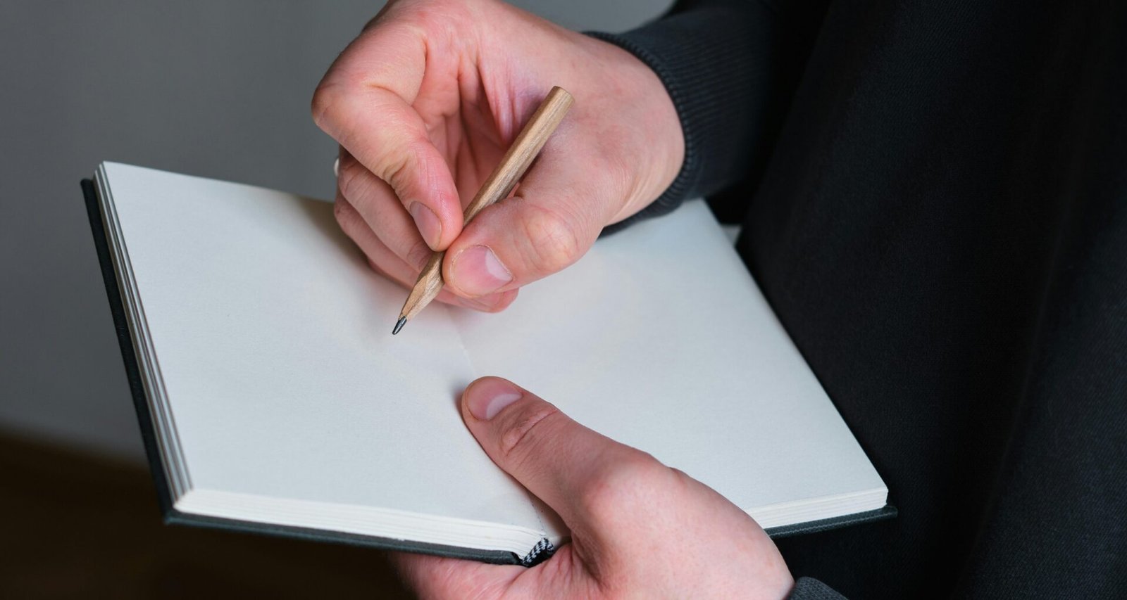 Person hält einen Bleistift und schreibt in ein geöffnetes leeres Notizbuch auf einem Schreibtisch