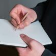 Person hält einen Bleistift und schreibt in ein leeres Notizbuch auf einem Schreibtisch