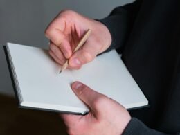 Person hält einen Bleistift und schreibt in ein leeres Notizbuch auf einem Schreibtisch
