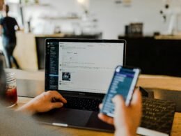 Person sitzt am Tisch und nutzt gleichzeitig ein Smartphone und einen Laptop zum Arbeiten.
