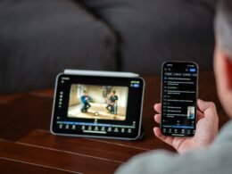 Eine Person sieht sich ein Video gleichzeitig auf einem Tablet und einem Smartphone an, Geräte liegen auf einem Holztisch.