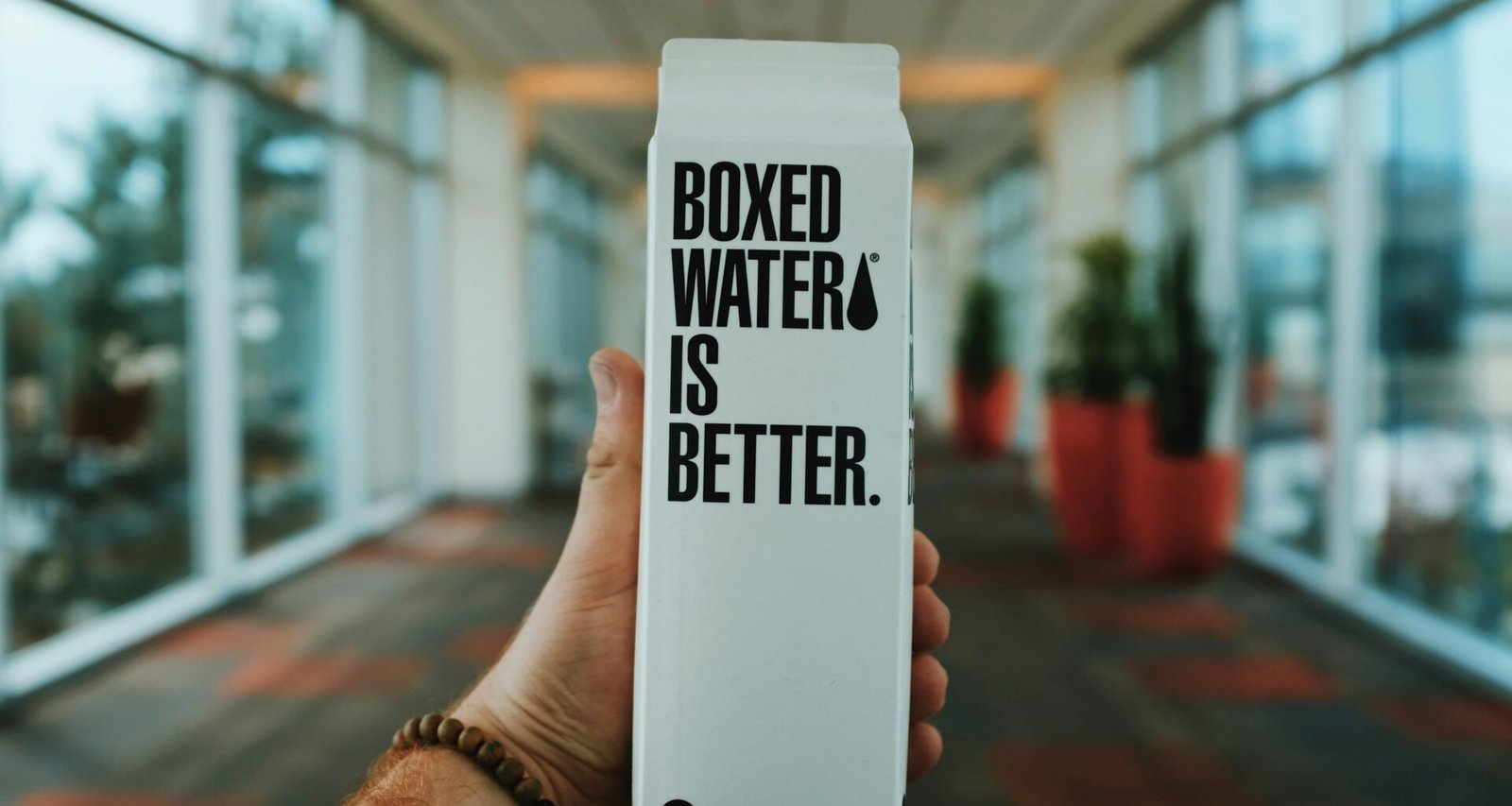 Eine Person hält eine Box mit der Aufschrift 'Boxed Water Is Better' in der Hand, Verpackung aus Karton als Alternative zu Plastik.
