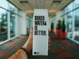 Eine Person hält eine Box mit der Aufschrift 'Boxed Water Is Better' in der Hand, Verpackung aus Karton als Alternative zu Plastik.