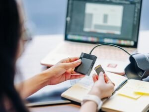 Person hält eine Speicherkarte in der Hand neben einem aufgeklappten Laptop und einem Notizbuch auf dem Schreibtisch.