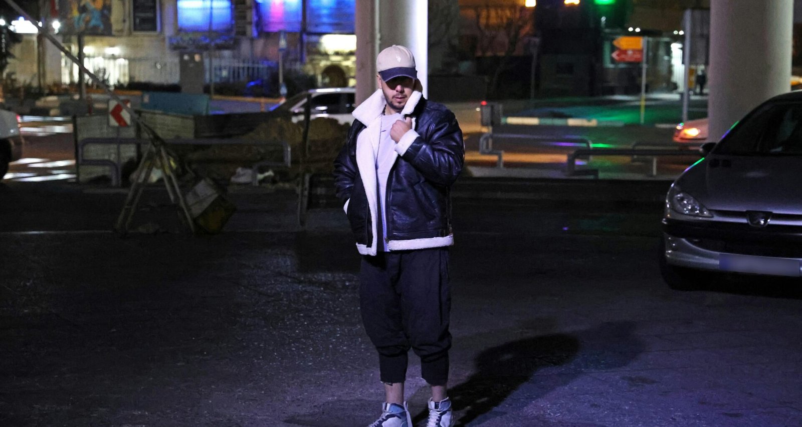 Eine Person in einer Jacke steht nachts auf einer belebten Straße, umgeben von Lichtern und urbaner Architektur.