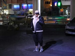 Eine Person in einer Jacke steht nachts auf einer belebten Straße, umgeben von Lichtern und urbaner Architektur.