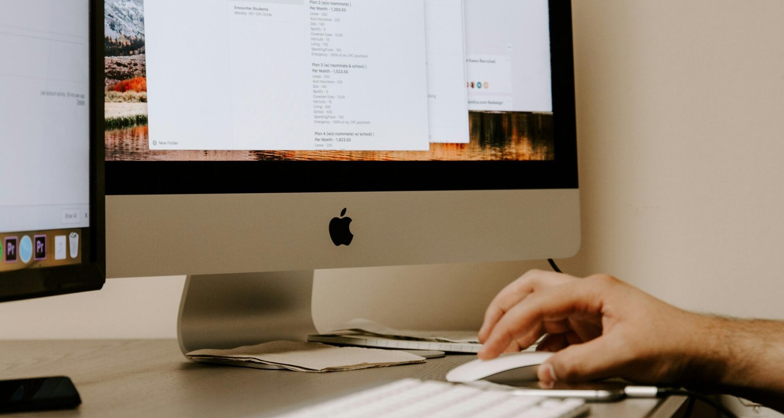Eine Person hält eine weiße Apple Magic Mouse neben einem iMac und einer Tastatur auf einem Schreibtisch.