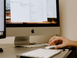 Eine Person hält eine weiße Apple Magic Mouse neben einem iMac und einer Tastatur auf einem Schreibtisch.