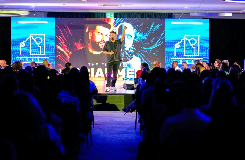 Ein Redner präsentiert auf einer Bühne vor einer sitzenden Zuhörerschaft während einer Konferenz.