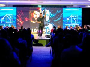 Ein Redner präsentiert auf einer Bühne vor einer sitzenden Zuhörerschaft während einer Konferenz.