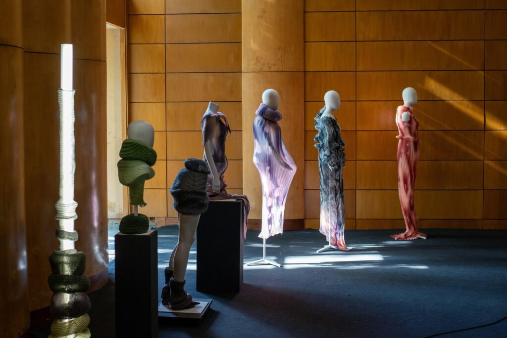 Mehrere Schaufensterpuppen mit abstrakten Skulpturen und ausgefallener Kleidung stehen in einem hellen Ausstellungsraum.