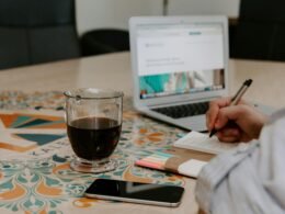 Person schreibt neben einem Glas Kaffee, einem ausgeschalteten iPhone und einem MacBook Air an einem Tisch