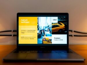 Ein aufgeklappter schwarzer Laptop zeigt einen Mann im gelben Hemd auf dem Bildschirm während einer Präsentation.