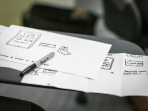 Ein schwarzer Marker liegt diagonal auf einem Stapel weißer Druckerpapiere und symbolisiert die Vorbereitung für Notizen oder Skizzen.