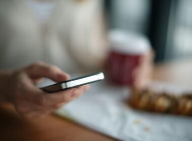 Person hält ein graues Smartphone in der Hand, Fokus liegt auf dem Gerät, im Hintergrund ist alles unscharf.