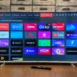 Smart TV Bildschirm mit verschiedenen App-Symbolen für Streaming, Spiele und Unterhaltung, fotografiert in moderner Wohnumgebung.