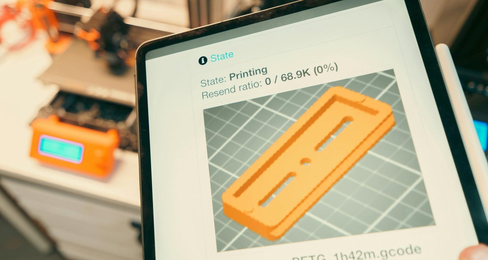 Tablet mit Anzeige des 3D-Druckfortschritts, im Hintergrund ein laufender 3D-Drucker in einer Werkstatt