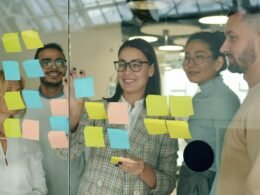 Mehrere Teammitglieder besprechen Konzepte und Ideen mit Haftnotizen auf einer Glaswand im modernen Büro.