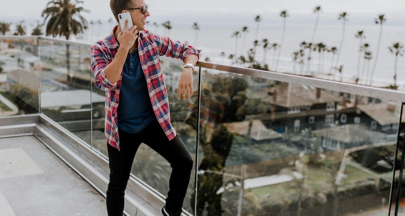 Person im roten Karohemd spricht auf einer Terrasse mit dem Handy; Holzgeländer im Hintergrund, Tageslicht