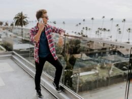 Person im roten Karohemd spricht auf einer Terrasse mit dem Handy; Holzgeländer im Hintergrund, Tageslicht