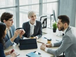 Mehrköpfiges diverses Business-Team arbeitet zusammen an einem großen Tisch in einem modernen Büro mit Laptops und Unterlagen.