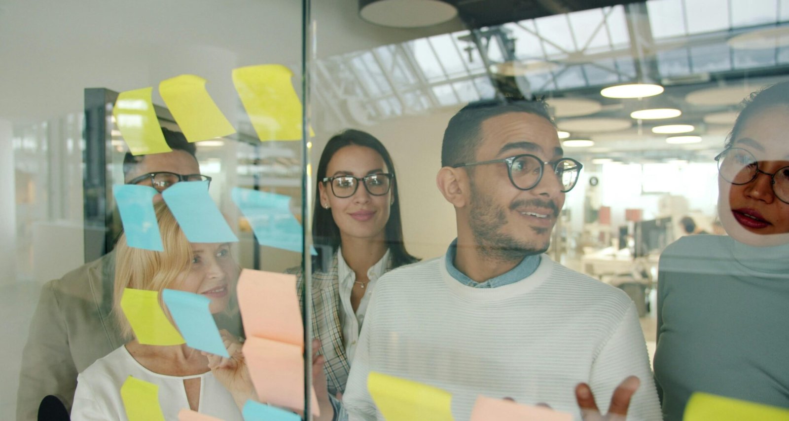 Diverse Teammitglieder brainstormen gemeinsam vor einer Glastafel mit bunten Haftnotizen im modernen Büro.
