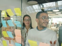 Diverse Teammitglieder brainstormen gemeinsam vor einer Glastafel mit bunten Haftnotizen im modernen Büro.